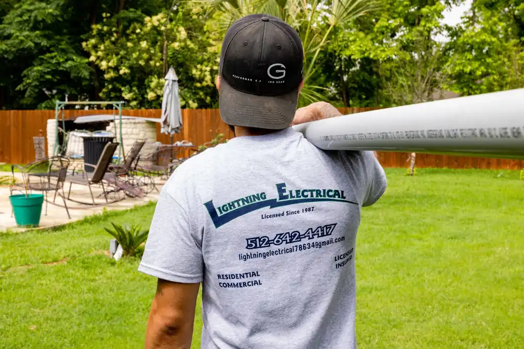 Technician seen from behind carrying PVC conduit at a residential backyard jobsite, wearing a Lightning Electrical branded shirt