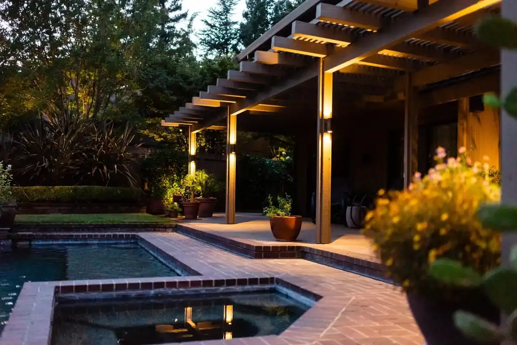 Twilight photo of an outdoor pergola and patio with integrated post lighting beside a pool