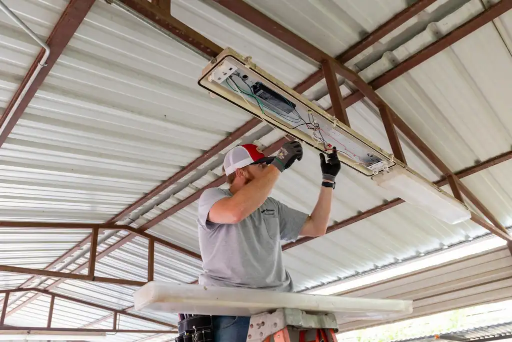 Technician wiring an overhead fluorescent fixture inside a metal roofed commercial structure
